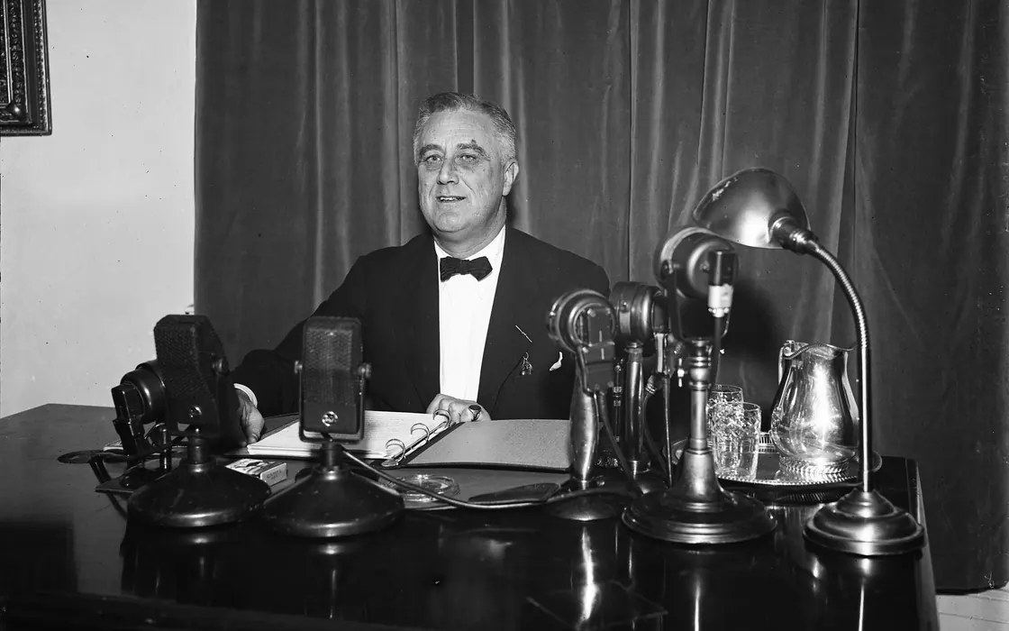 Fotografia de 1937: Franklin D. Roosevelt sentado a uma escrivaninha, falando diante de cinco microfones de rádio enfileirados, com um caderno aberto à frente e uma cortina pesada ao fundo