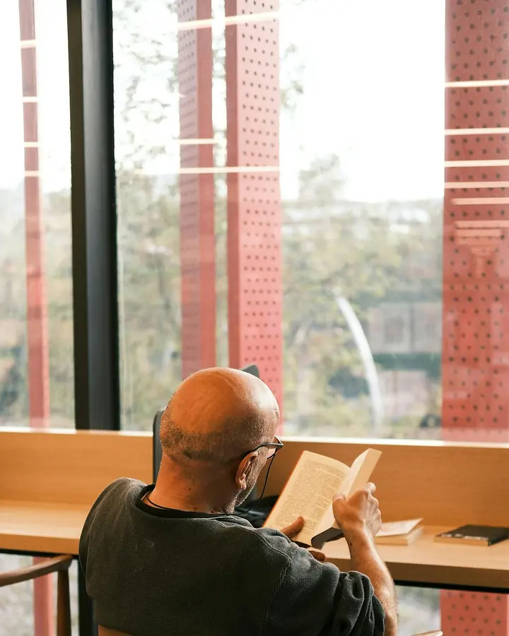 Homem sentado numa biblioteca, de costas, lendo um livro diante de uma janela ampla