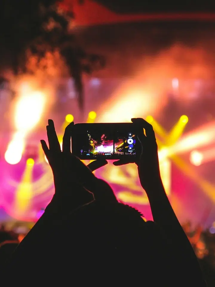 Mãos erguidas segurando um celular que filma o palco de um show, com luzes coloridas ao fundo.